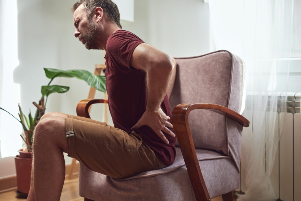 Older man sitting in chair and holding his hip in pain and in need of surgery to correct adult hip dysplasia